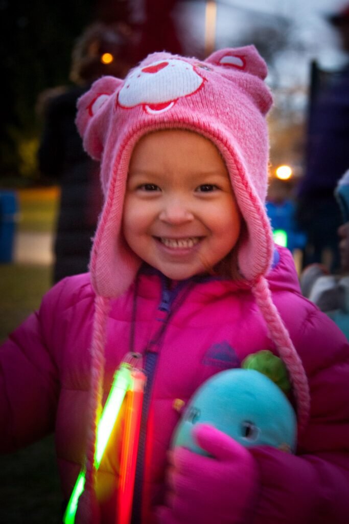 A smiling child wears a pink bear hat and jacket while holding a blue plush toy with glowing lights.
