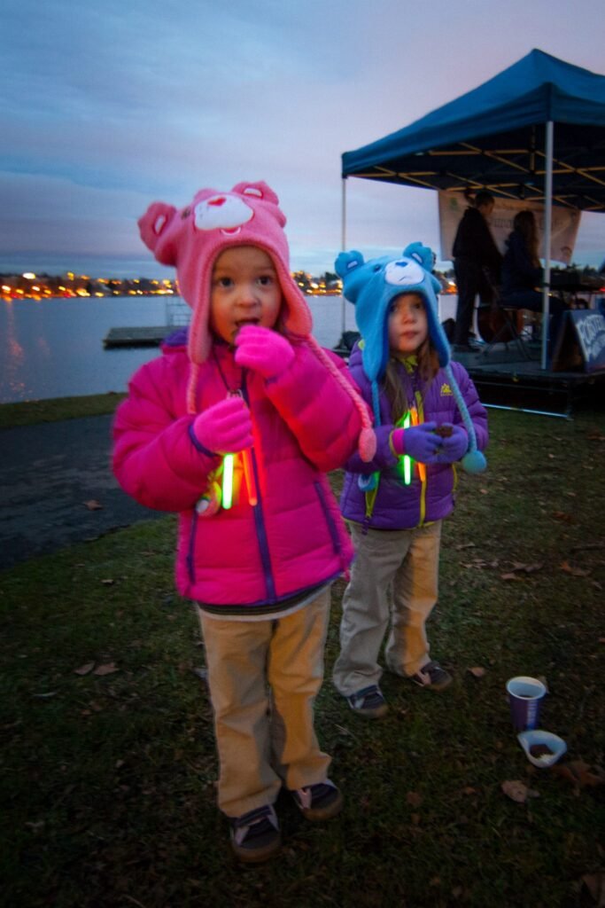 Children wearing cozy bear hoods enjoy glowing lanterns at a lakeside event in the Pacific Northwest.