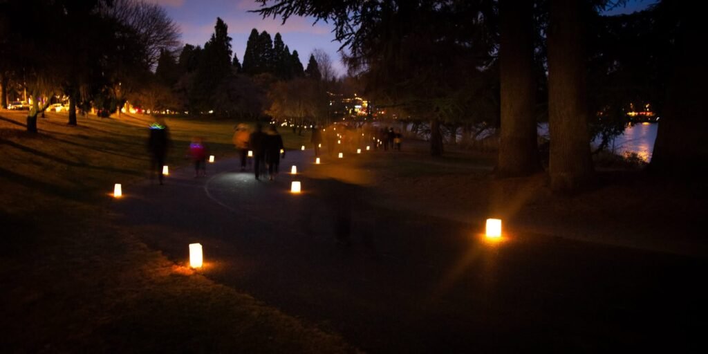 A group of people walk a park path lined with glowing lanterns at dusk.