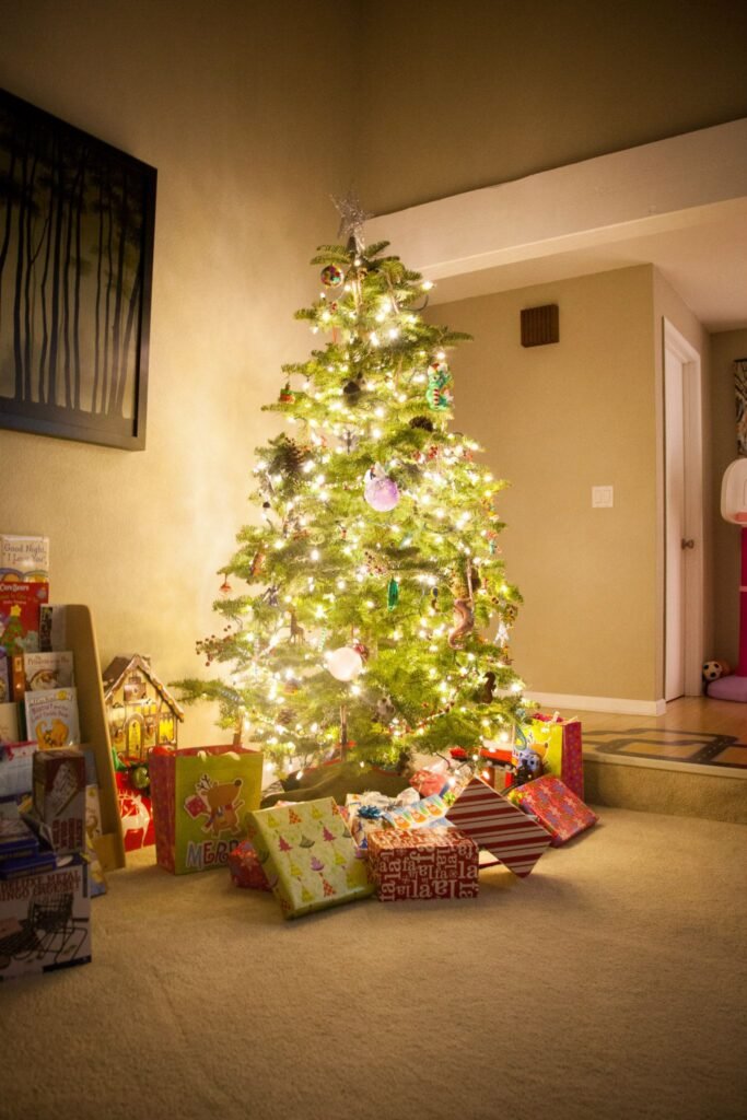 A warmly lit Christmas tree decorated with white lights and ornaments stands in a home living room with wrapped gifts beneath it.