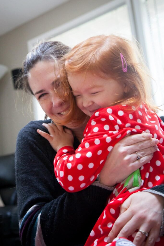 An adult and a young child in pink polka dot pajamas share a close, cheerful hug near a window.