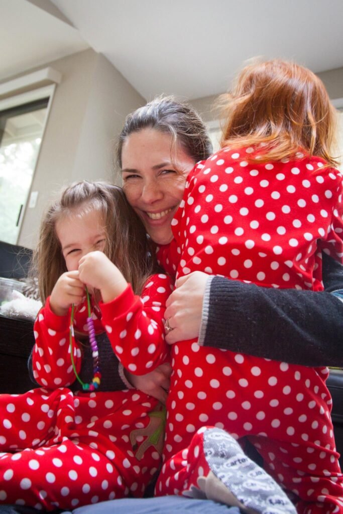 A woman smiles as she hugs two children wearing matching red and white polka dot pajamas. Both children are seated while covered in festive holiday pajamas.