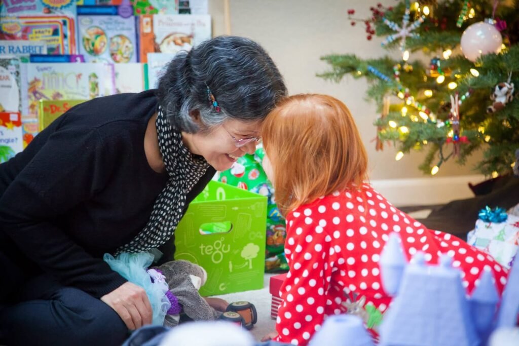 Two people share a joyful moment by a decorated Christmas tree with books in the background. Festive lights and holiday decorations create a cozy atmosphere.