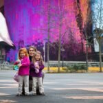 A family poses against a vivid mural at a modern plaza in Vancouver's Olympic Village, surrounded by bare trees and clear skies.