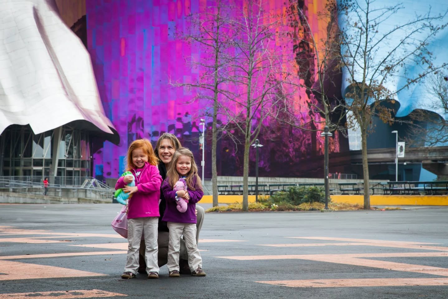 A family poses against a vivid mural at a modern plaza in Vancouver's Olympic Village, surrounded by bare trees and clear skies.