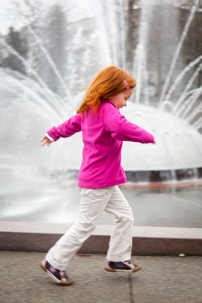 A child in a pink jacket walking near a large public fountain with water jets spraying upwards.