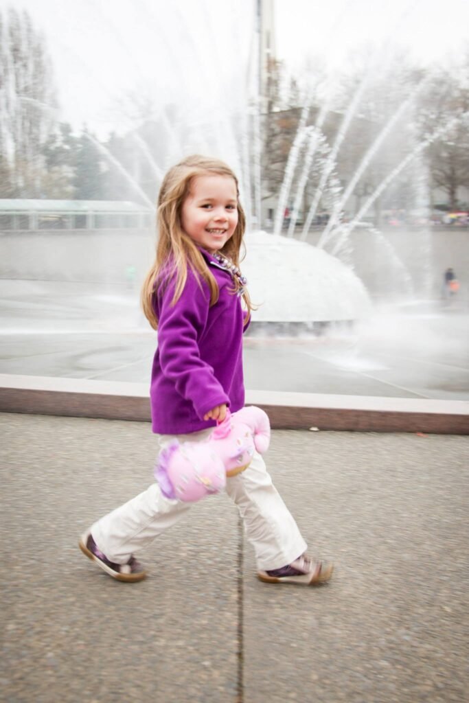 A young child in a purple fleece jacket walks smiling in front of a large water fountain.