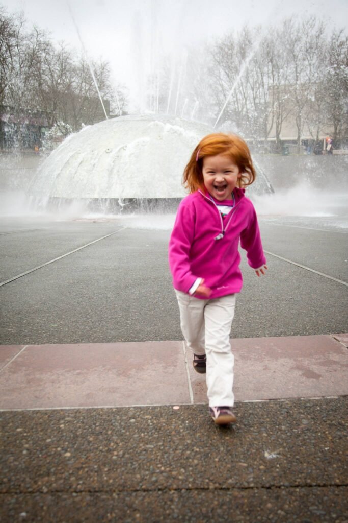 A smiling child in a pink jacket runs near a large public fountain emitting mist. The background features bare winter trees and paved walkways typical of a park setting in the Pacific Northwest.