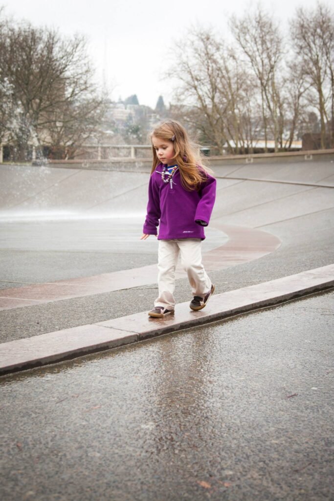 A child wearing a purple jacket walks on a wet walkway at a park on a cloudy day with leafless trees in the background.
