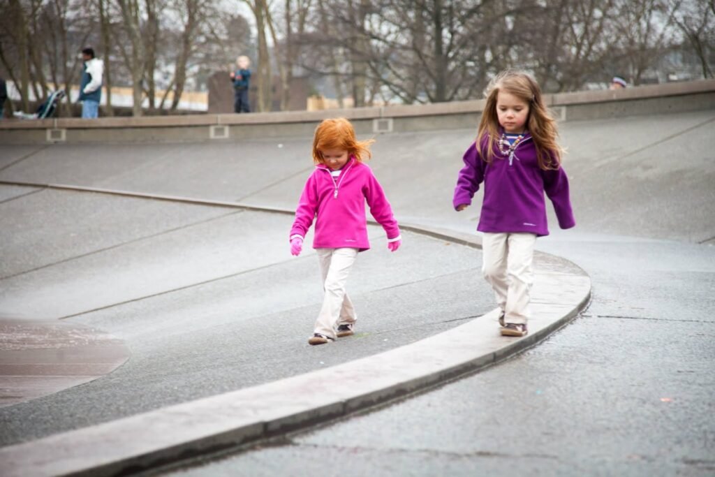 Two young girls walking through a park on a cool day, wearing pink and purple jackets.