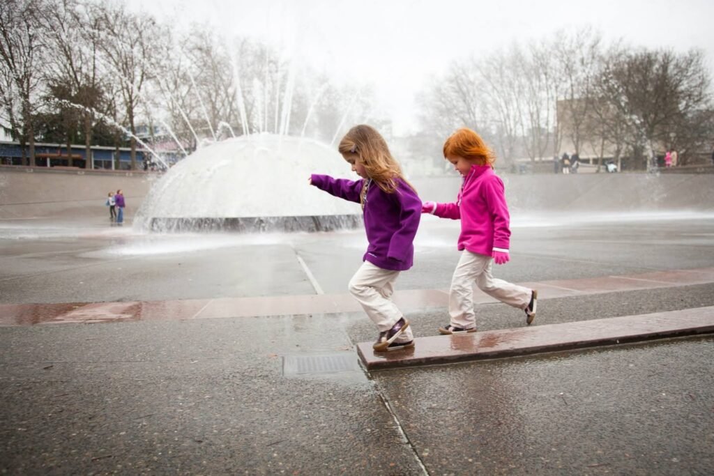 Two children in colorful jackets balance on a ledge near a large fountain during a cloudy winter day.