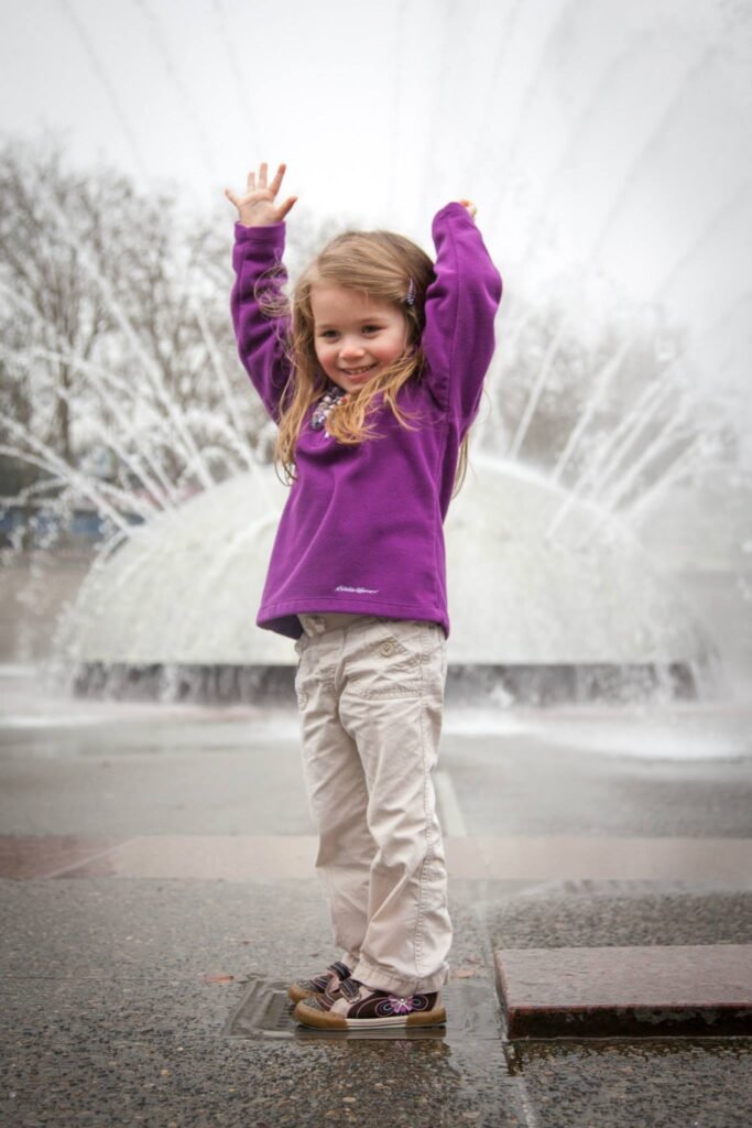 A child in a purple sweater waves with arms raised in front of a large city fountain with a blurred background.