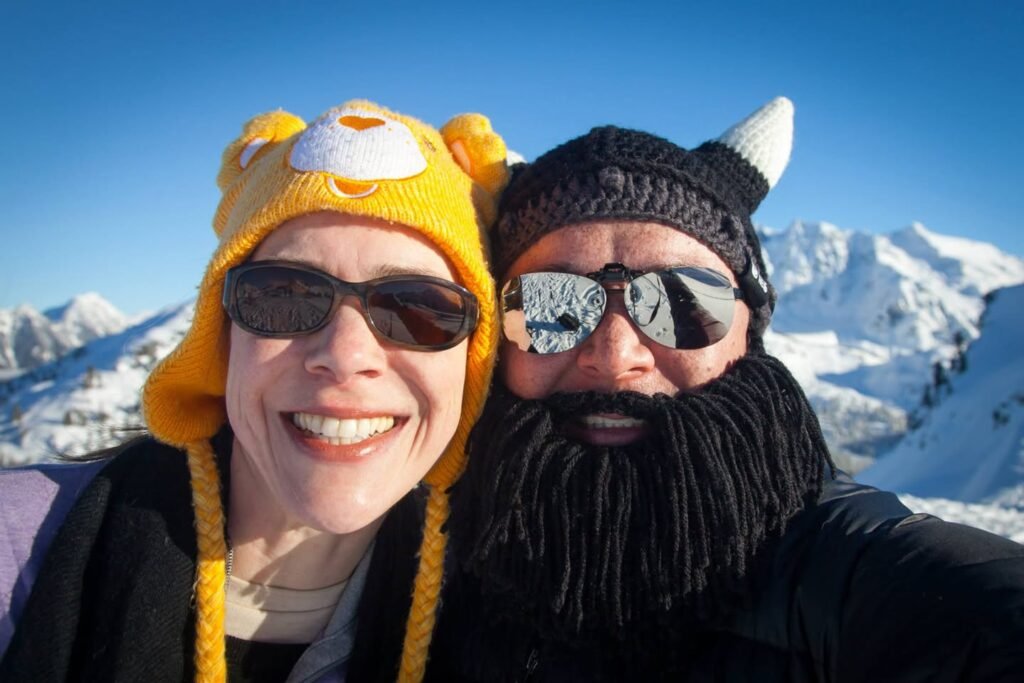A couple in colorful knit hats takes a selfie against a backdrop of snow-covered mountains and blue sky.