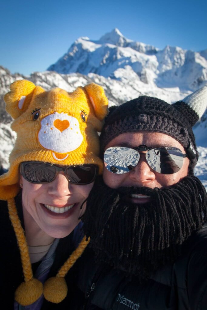 Two people wearing sunglasses and colorful hats in front of snow-covered mountains.