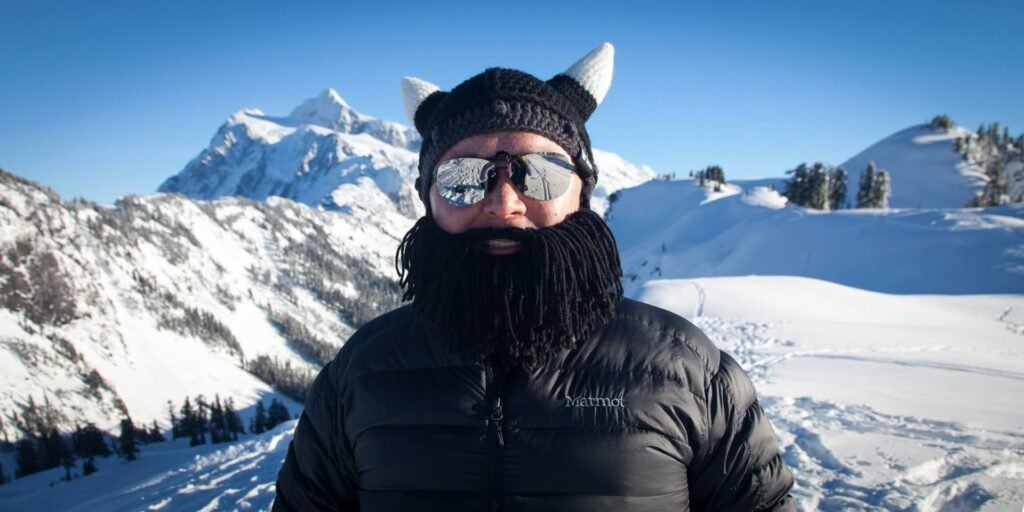 A bearded individual wearing a hat with cat ears and sunglasses stands before a snow-covered mountain range. They are dressed in a black puffer jacket, enjoying a winter mountain view.