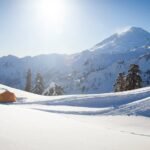 A winter mountain landscape with snow-covered peaks, tall evergreens, and a bright sun shining in a clear blue sky.