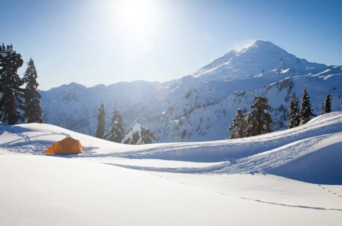 A winter mountain landscape with snow-covered peaks, tall evergreens, and a bright sun shining in a clear blue sky.