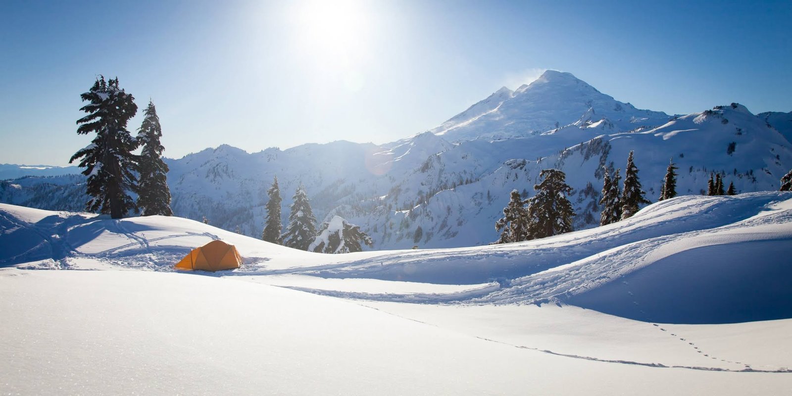 A winter mountain landscape with snow-covered peaks, tall evergreens, and a bright sun shining in a clear blue sky.