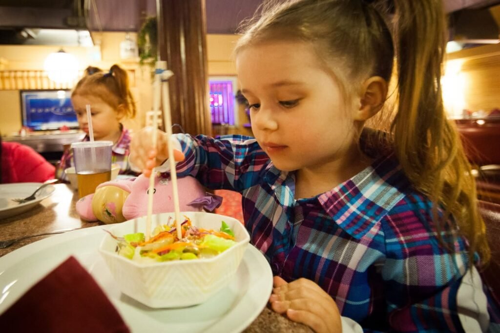 A child using chopsticks to eat a bowl of food in a warm, inviting restaurant setting. Another child enjoys a drink at the table.
