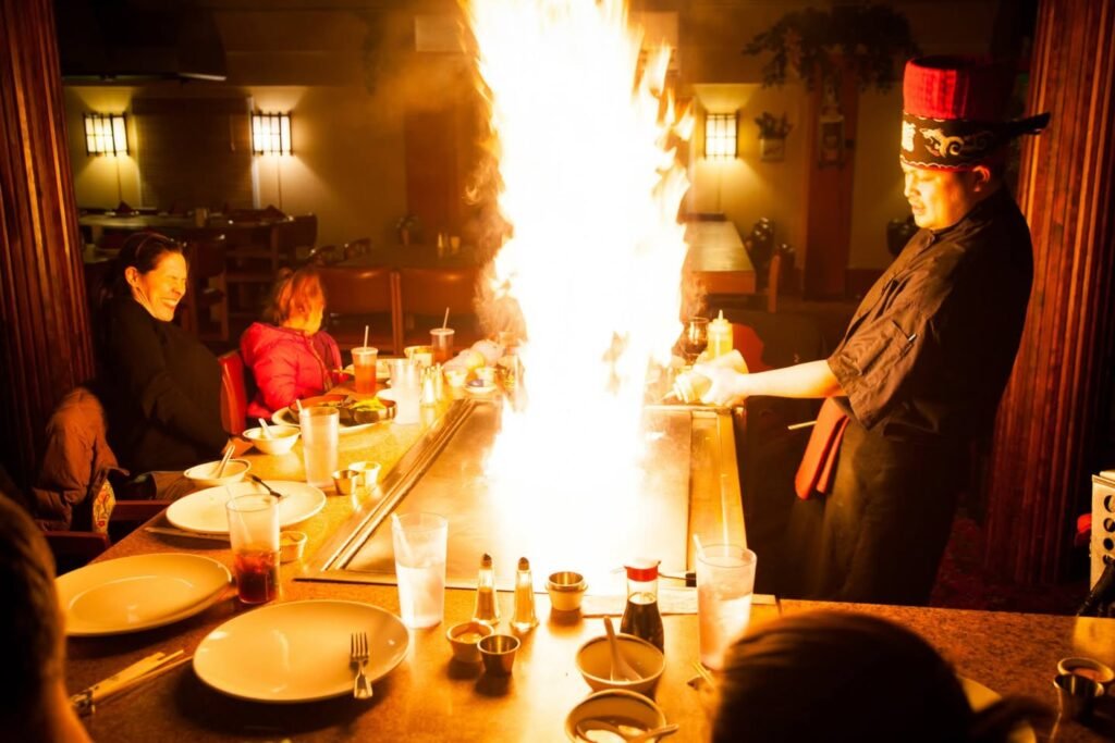 A server performs a flambé at a sushi bar, creating a large flame over the counter. Diners watch the culinary show in a warmly lit dining room.
