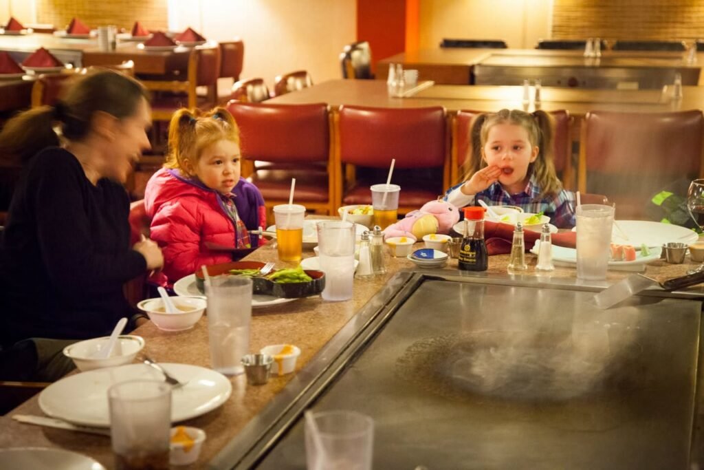 A family enjoying a teppanyaki dinner at a restaurant with a large cooking grill and steam rising from the iron surface. Several children and adults are seated along the counter, watching food preparation.