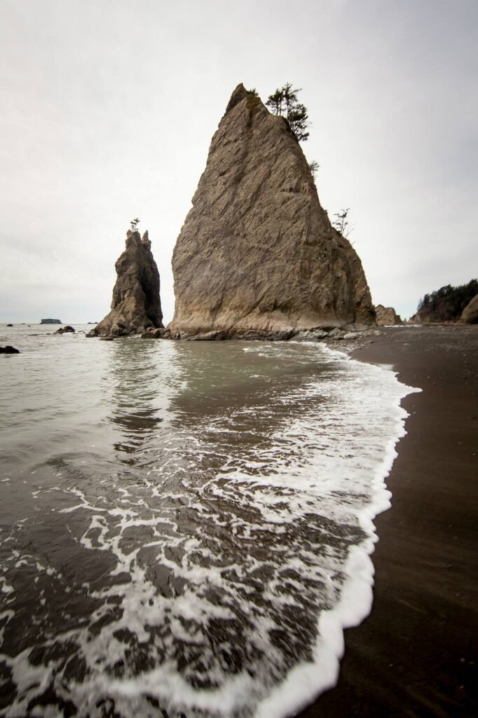 Towering seastacks rising from grey waters with crashing waves breaking on a sandy beach.