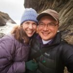 A couple enjoying a sunny day at rugged coastal cliffs, bundled up in winter hats and jackets.