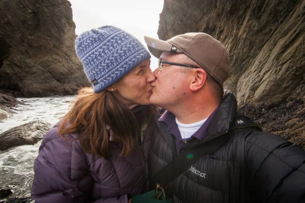 A couple shares a tender kiss in front of dramatic coastal rocks.