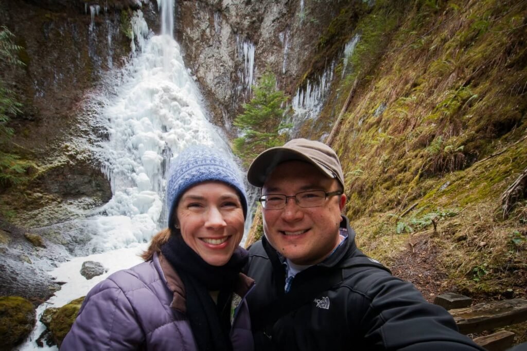A couple poses for a selfie in front of a large, cascading waterfall. They are dressed in warm winter gear, including hats and jackets, set against the rugged landscape.