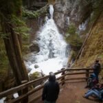 Rialto Beach People standing on a wooden platform overlooking a powerful waterfall cascading down a rocky cliff in the Pacific Northwest.