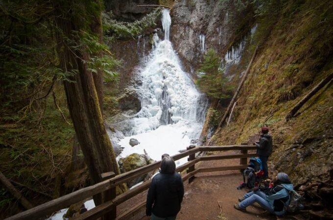 People standing on a wooden platform overlooking a powerful waterfall cascading down a rocky cliff in the Pacific Northwest.