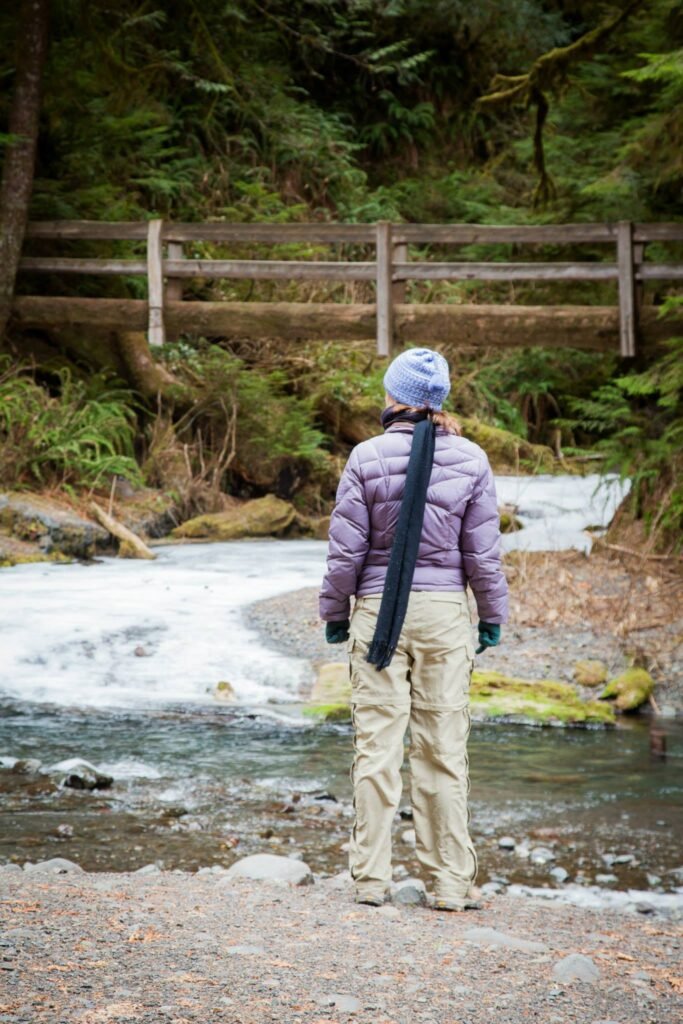 A person with a camera strap stands by a stream in a forested area, with a wooden fence and lush vegetation in the background.