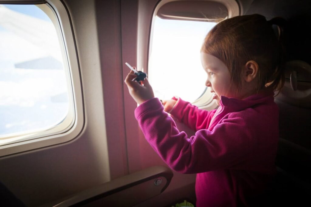 A young passenger in a pink fleece jacket looks out an airplane window at a wing and holds a small object.