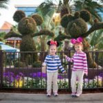 Two children in striped shirts and headbands pose with topiary sculptures and flowers at a theme park. The vibrant purple flowers add color to the cheerful outdoor scene.