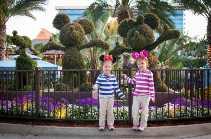 Two children in striped shirts and headbands pose with topiary sculptures and flowers at a theme park. The vibrant purple flowers add color to the cheerful outdoor scene.