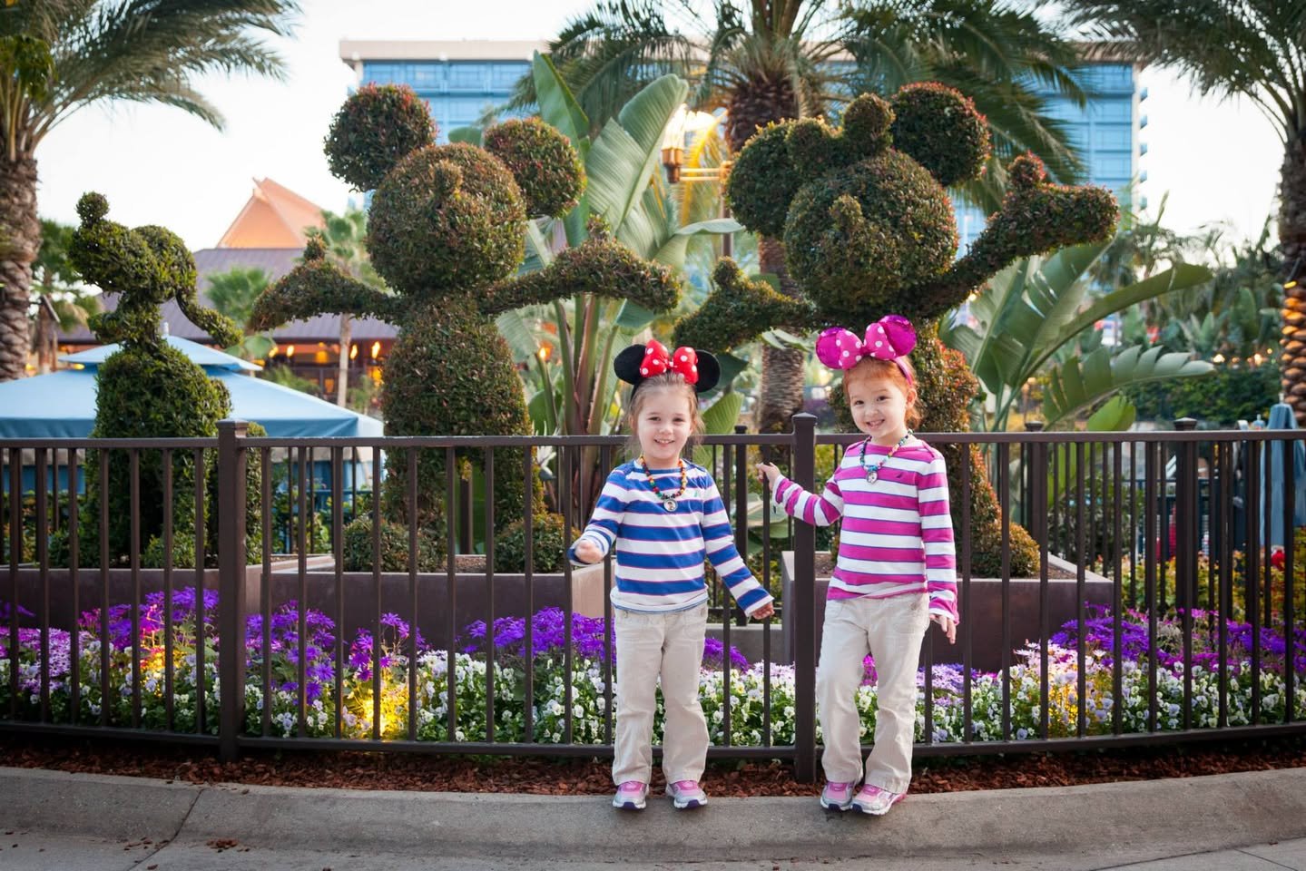 Disneyland! Days 1 & 2 Two children in striped shirts and headbands pose with topiary sculptures and flowers at a theme park. The vibrant purple flowers add color to the cheerful outdoor scene.