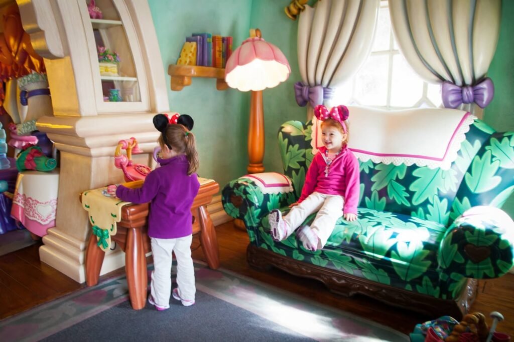 Two children wearing Disney ear headbands play in a colorful themed room with a patterned sofa. The scene features a white fireplace, a glowing lamp, and decorative curtains.