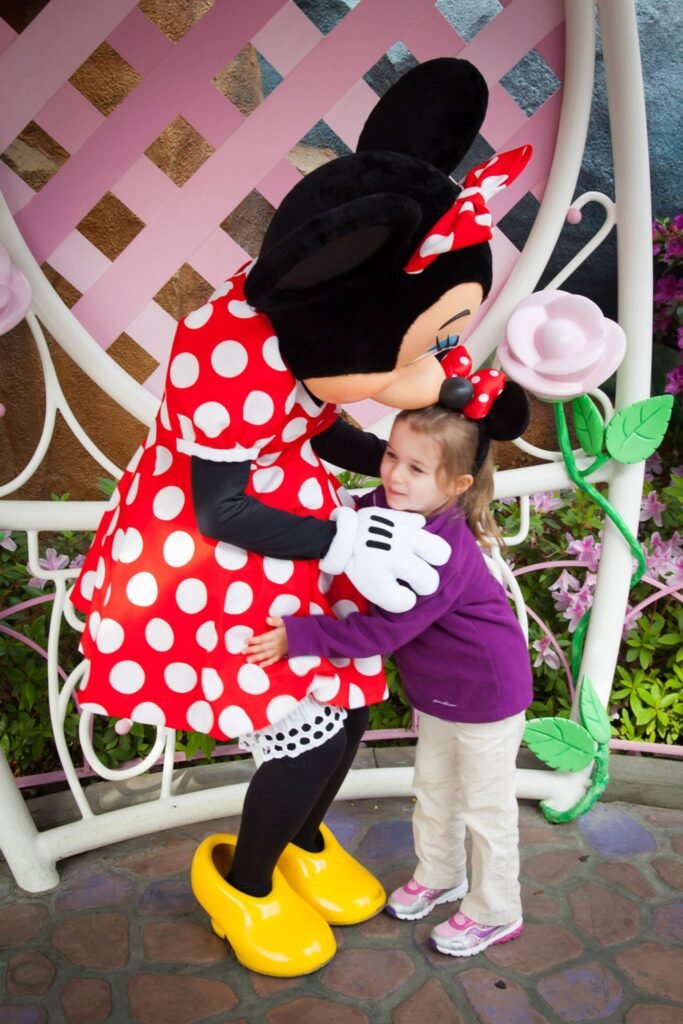 A child hugs Minnie Mouse in front of a pink lattice wall with a decorative rose.