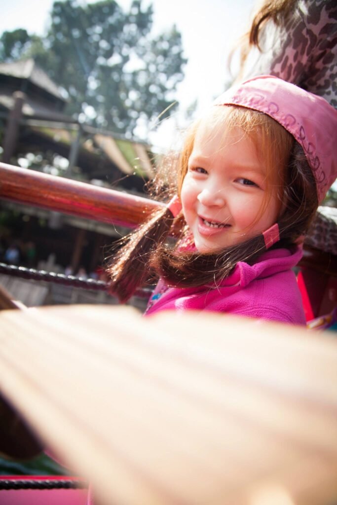Close-up of a smiling person wearing a pink hat and scarf. Background shows trees and architectural elements.