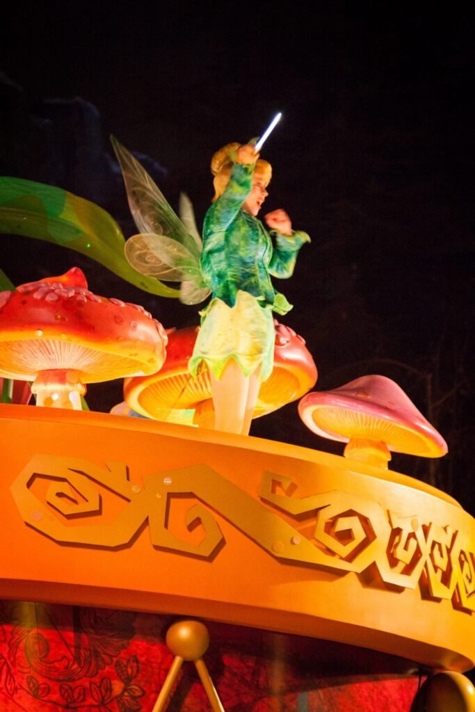 A glowing Tinker Bell figure stands atop a vibrant carousel float with mushroom decorations, captured against a dark night sky in a lively parade scene.