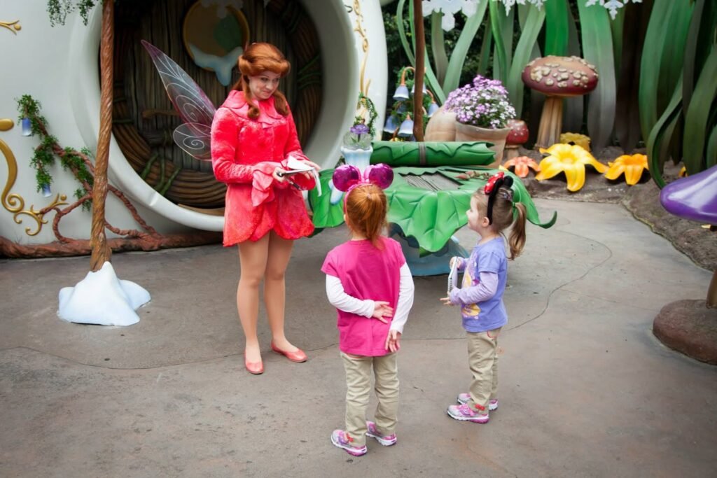 A fairy garden setting with whimsical decorations and a child wearing pink Mouse Ears visiting a character in red attire.