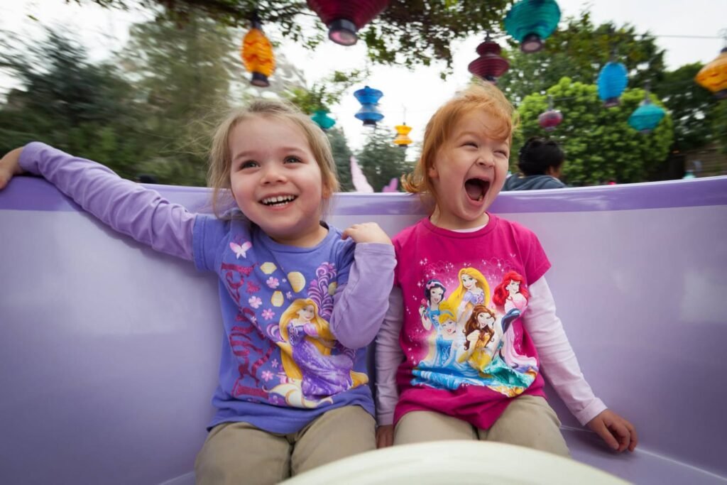 Two children ride a purple coaster under colorful hanging decorations with trees in the background. This photo captures the excitement of a family day at the amusement park.