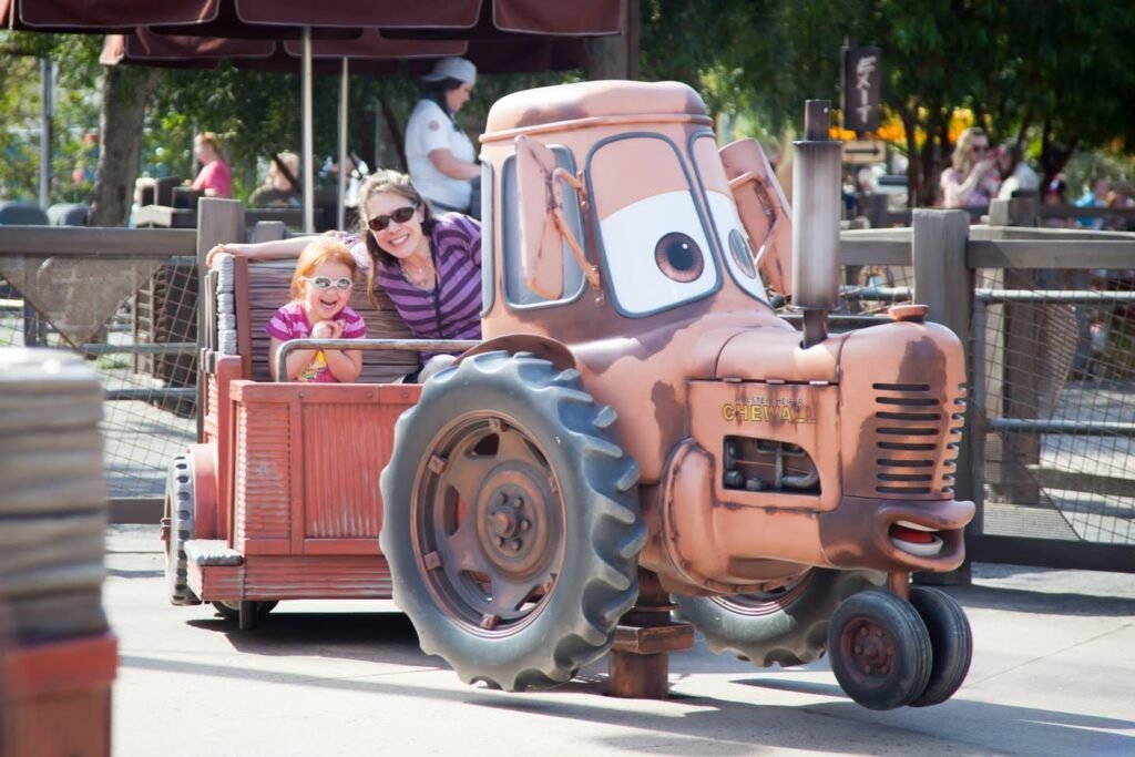 A smiling woman and child are riding in a red farmhouse wagon pulled by a character tractor. The scene captures a magical moment at a theme park during daylight.