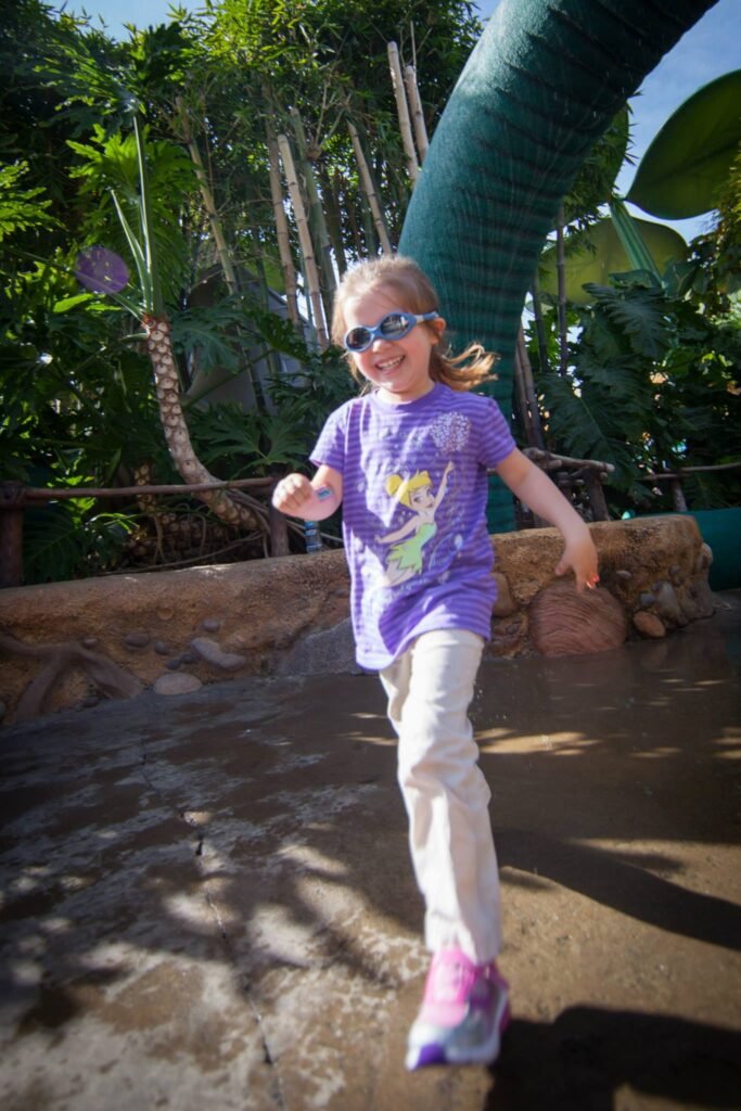 A child wearing a purple t-shirt and white pants walks on a wet stone path near a large water slide and lush green foliage.