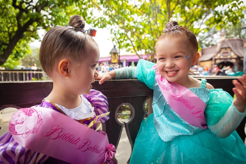 Two young children wearing princess costumes and sashes smile on a park ride. One child wears a teal dress with a face marking, while the other wears purple with a princess sash.