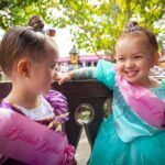 Two young children wearing princess costumes and sashes smile on a park ride. One child wears a teal dress with a face marking, while the other wears purple with a princess sash.