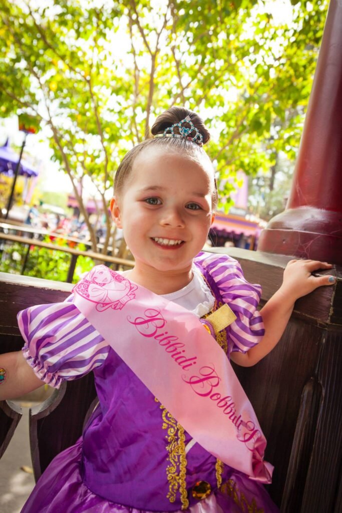 A young person in a purple Belle princess costume and sash enjoys a sunny day at the park.
