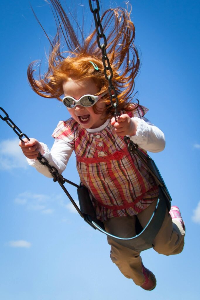 A child swinging high on a chain swing with hair blowing in the wind under a bright blue sky. Wide-angle and motion shots capture the fun of a summer day outdoors.