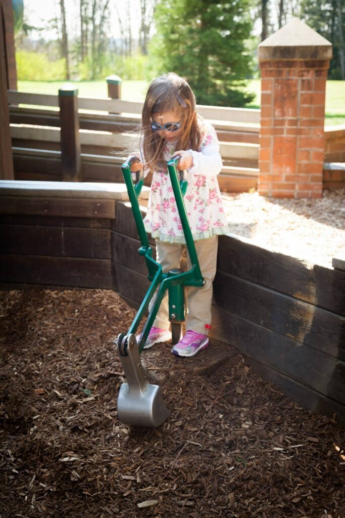 A child uses a green garden tool in a dirt pit near a brick structure.
