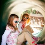 Two children wearing sunglasses sitting inside a green playground tunnel. They appear to be enjoying their time at a playground.